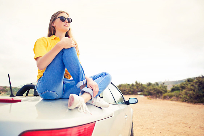 Valkeakosken ammattiopisto, VAAO. Henkilö istuu auton päällä. Hänellä on aurinkolasit, keltainen paita, siniset farkut ja valkoiset kengät päällä.