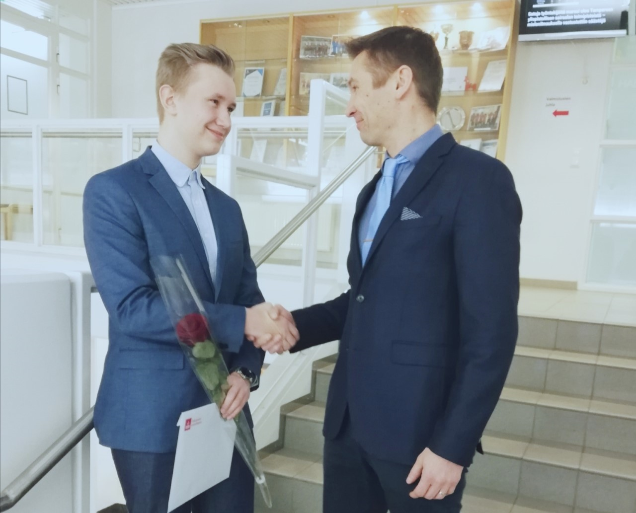Valkeakosken ammattiopisto, VAAO. Toimialapäällikkö Mikko Siirilä kättelee portaikossa valmistunutta oppilasta. Oppilaalla on kädessään punainen ruusu ja VAAOn kirjekuori.