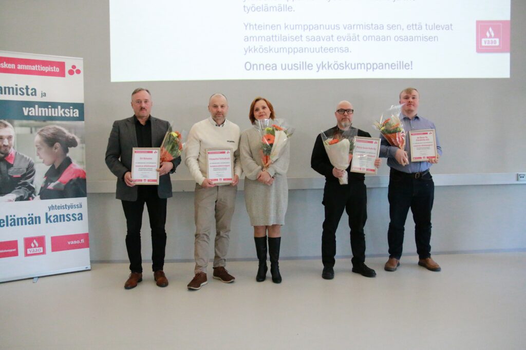 Viisi henkilöä seisoo rivissä, kukkakimput ja diplomit kädessä.