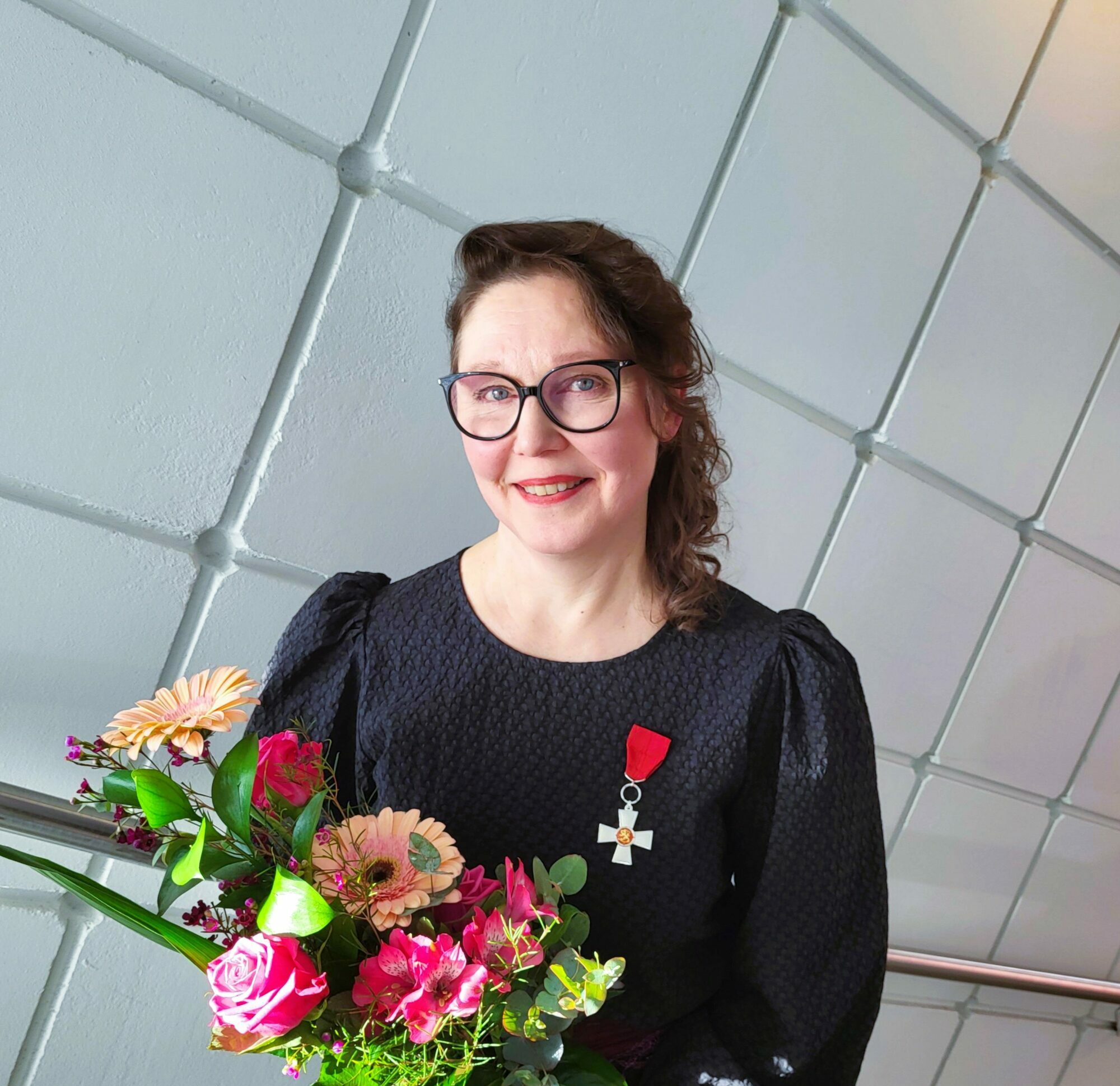 Tummahiuksinen, henkilö hymyilee kukkakimppu kädessä ja ansiomerkki rinnassa. Silmälasit, tumma puku