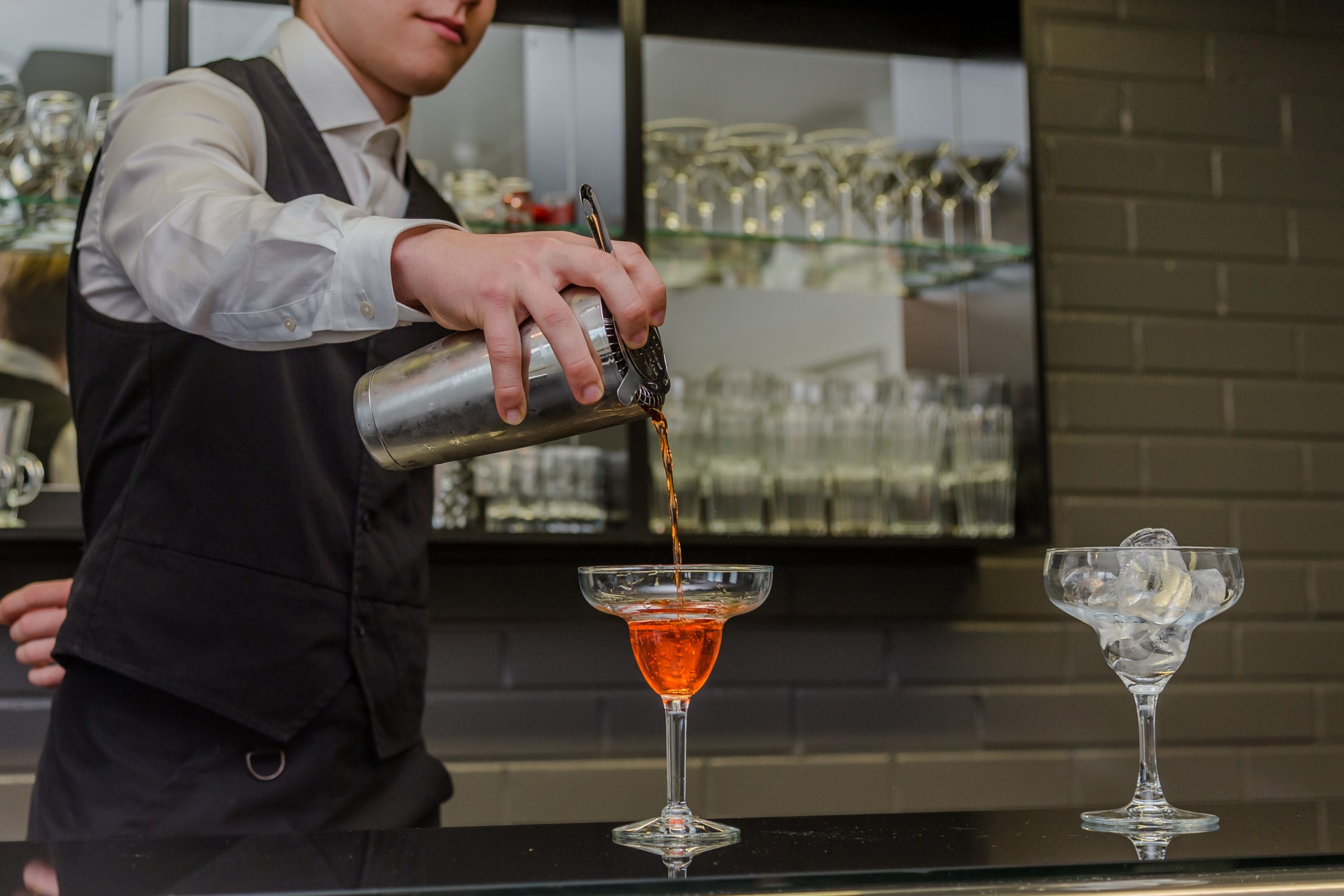 Valkeakosken ammattiopisto, VAAO, ravintolatyöntekijän työvaatteisiin pukeutunut henkilö annostelee punaista drinkkiä baaritiskillä olevaan korkeajalkaiseen lasiin.