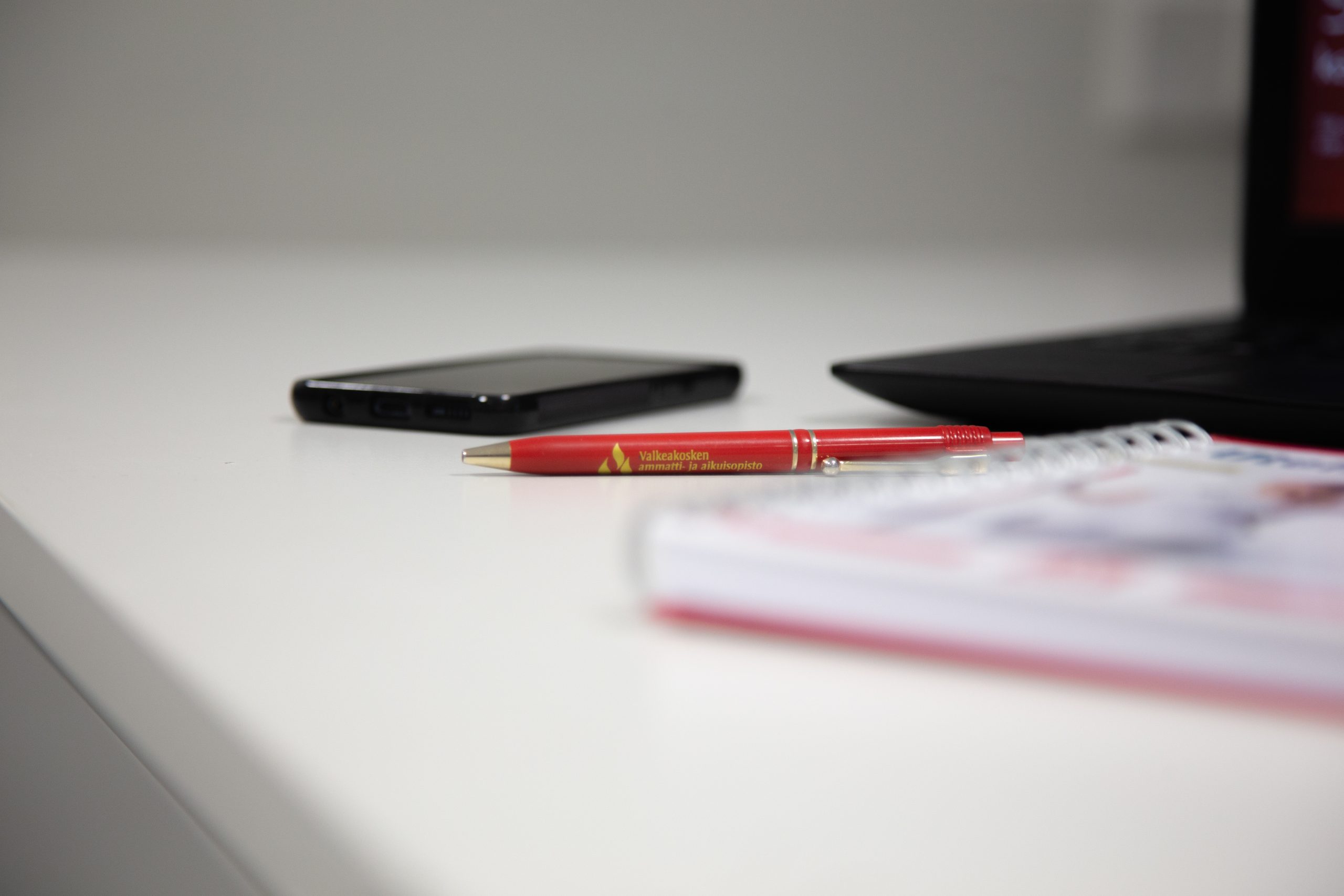 Valkeakosken ammattiopisto, VAAO. Kuvassa valkoisella pöydällä näemme mustan älypuhelimen, VAAOn-punaisen kuulakärkikynän, vaalean kierrevihon ja mustan kannettavan tietokoneen kulmaa.