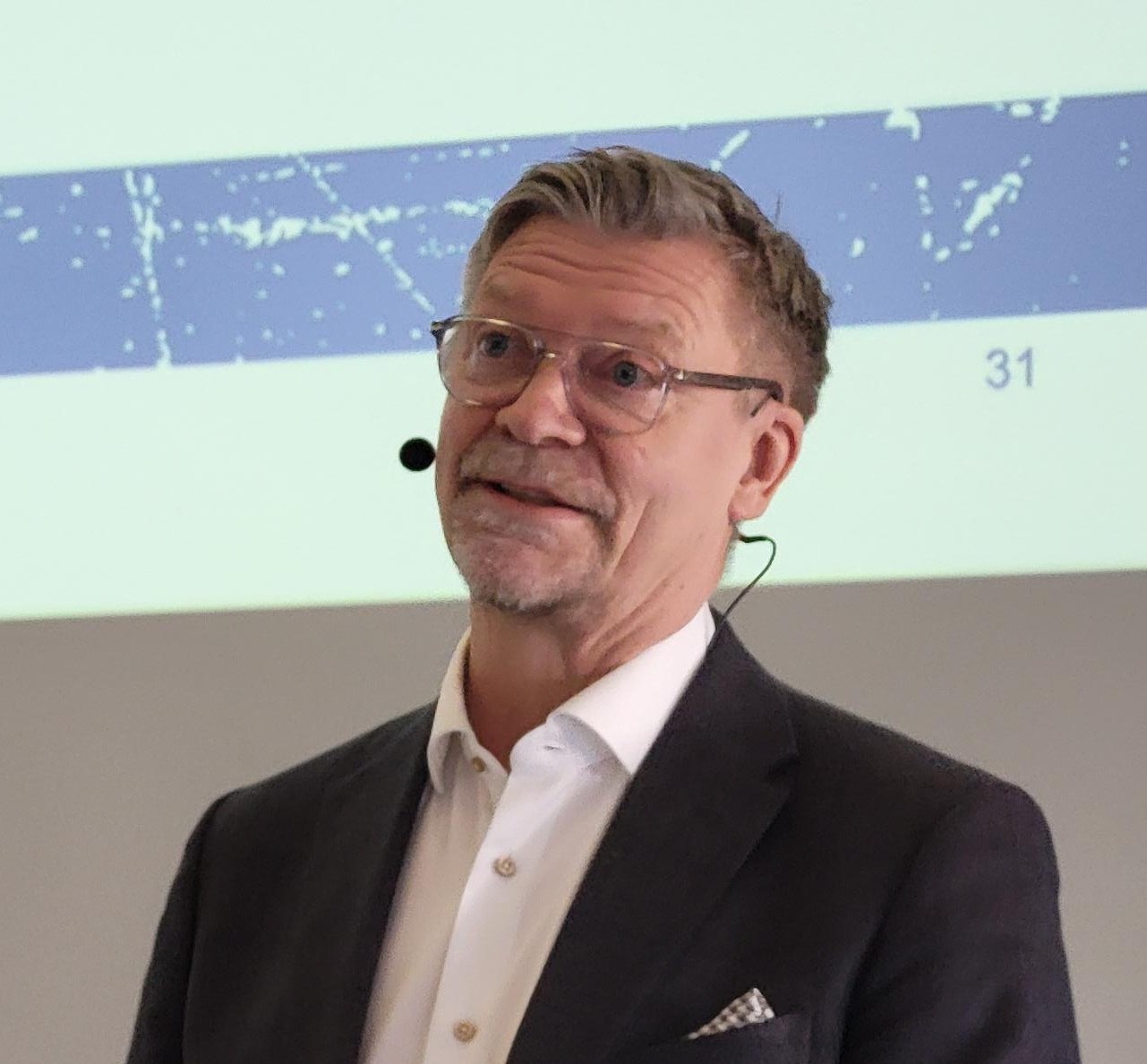 Jääkiekkovalmentaja Jukka Jalonen pitää esitystä tiimityöstä. Tumma puku päällä, valkoinen kauluspaita, silmälasit.