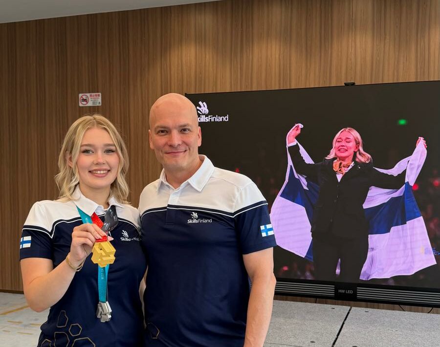 Kultamitalin voittanut kilpailija World Skills Sanghai2026-kisojen jälkeen seisoo valmentajansa kanssa screenin edessä, jolta näkyy kilpailijan kuva ja Suomen lippu