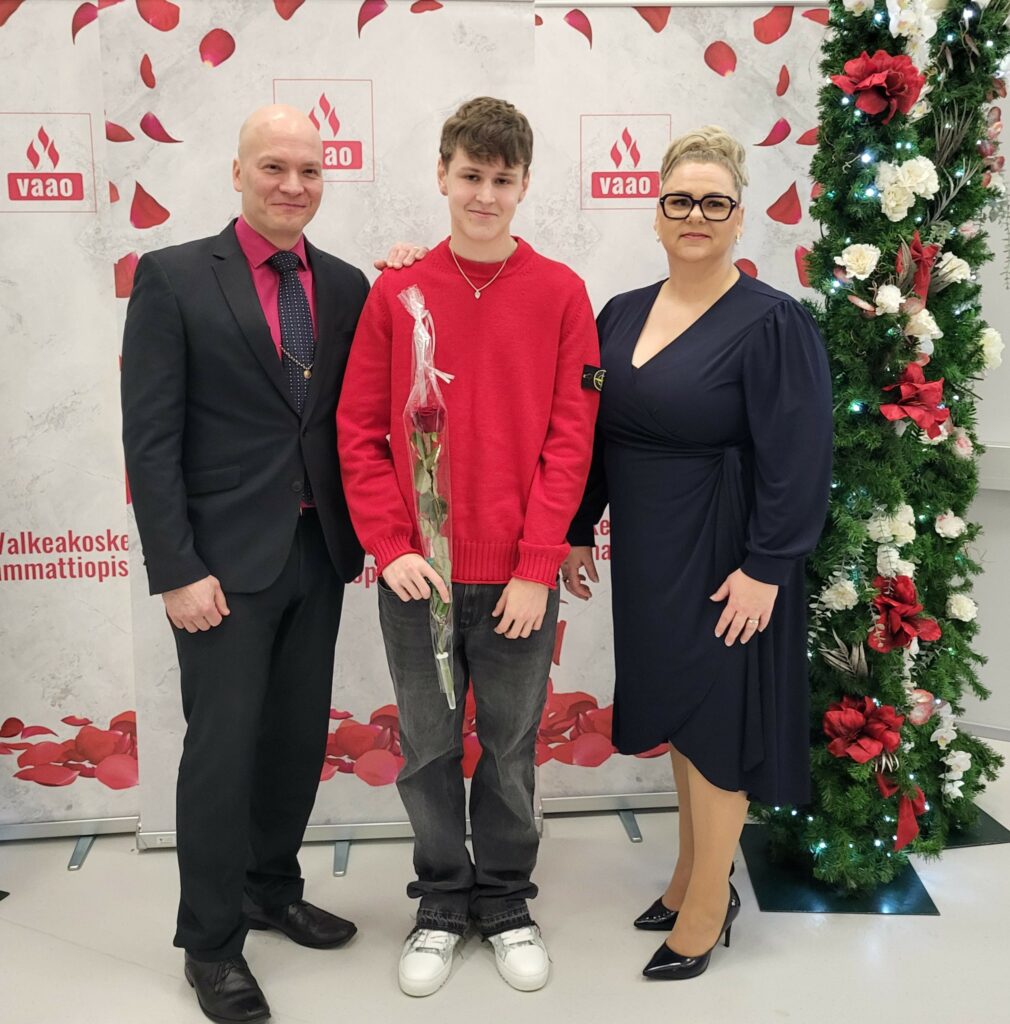 Valkeakosken ammattiopisto, VAAO, Kolme henkilöä seisoo kuvausseinän edessä ja hymyilee kameralle, keskimmäisellä on yksi punainen ruusu kädessä.