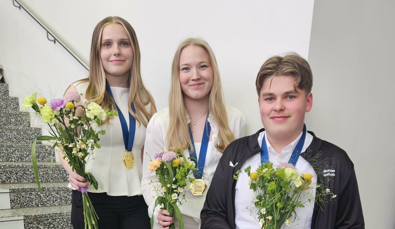 Kolme opiskelijaa seisoo vierekkäin, kukkakimput käsissä, hymyilevät kameralle