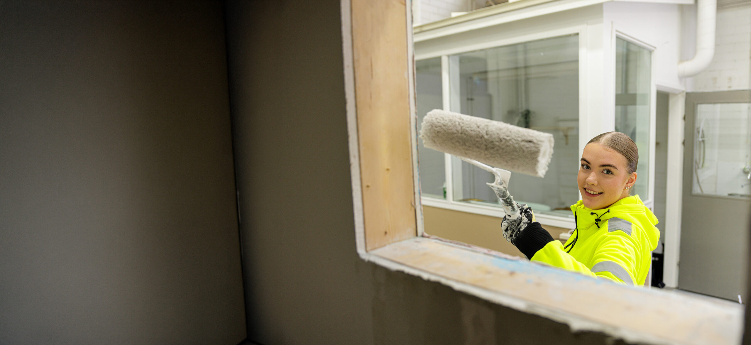 VAAOssa pintakäsittelyä opiskeleva tuleva maalari maalaa seinää maalitelalla. Kuva otettu toiselta puolelta. Opiskelija kurkkaa ikkunasta kohti valokuvaajaa.