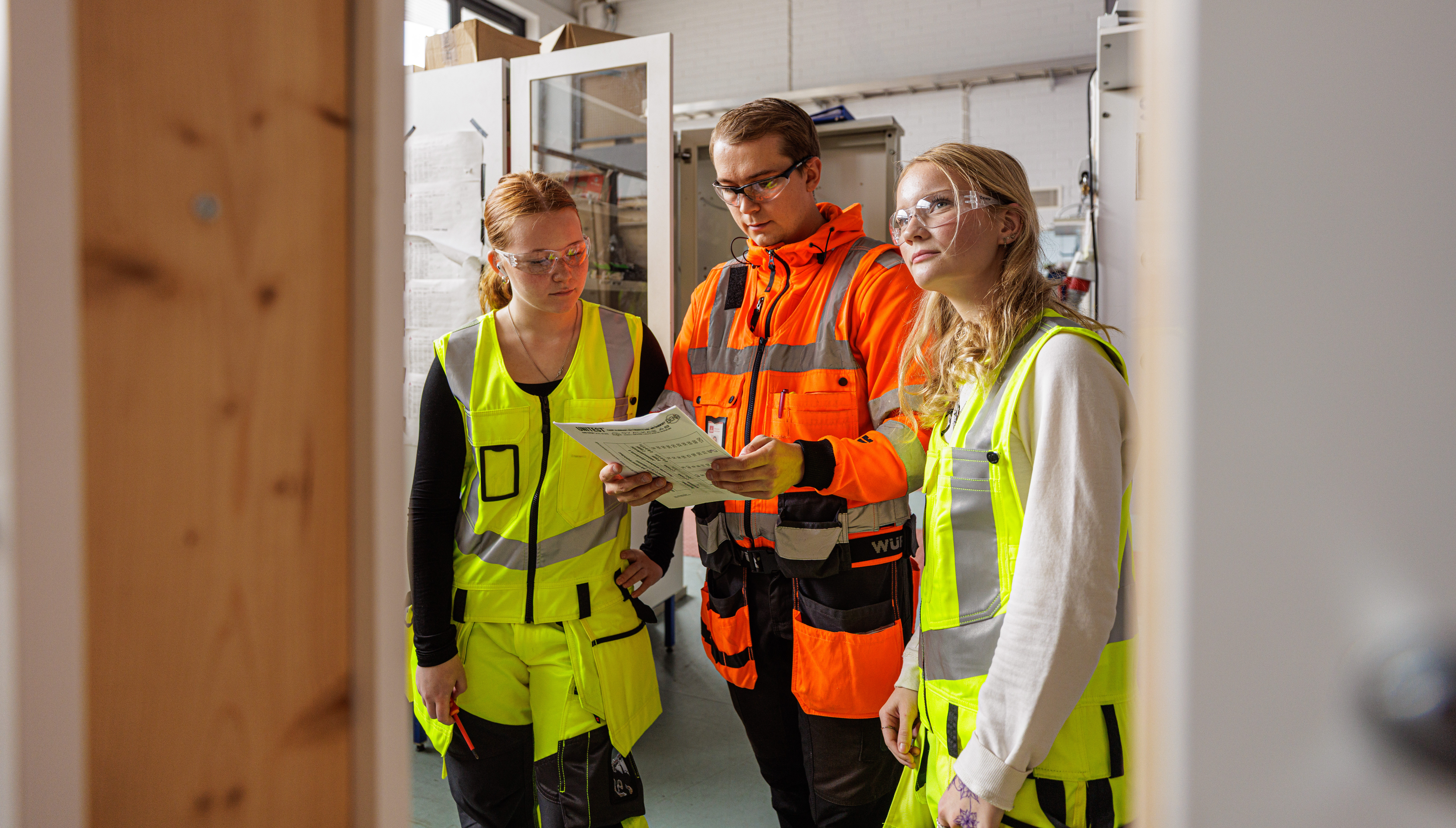 Opettaja seisoo keskellä, kuvassa kaksi opiskelijaa opettajan kanssa. Kaikilla työvaatteet ja suojavarusteet päällä. Katsovat työohjetta yhdessä.