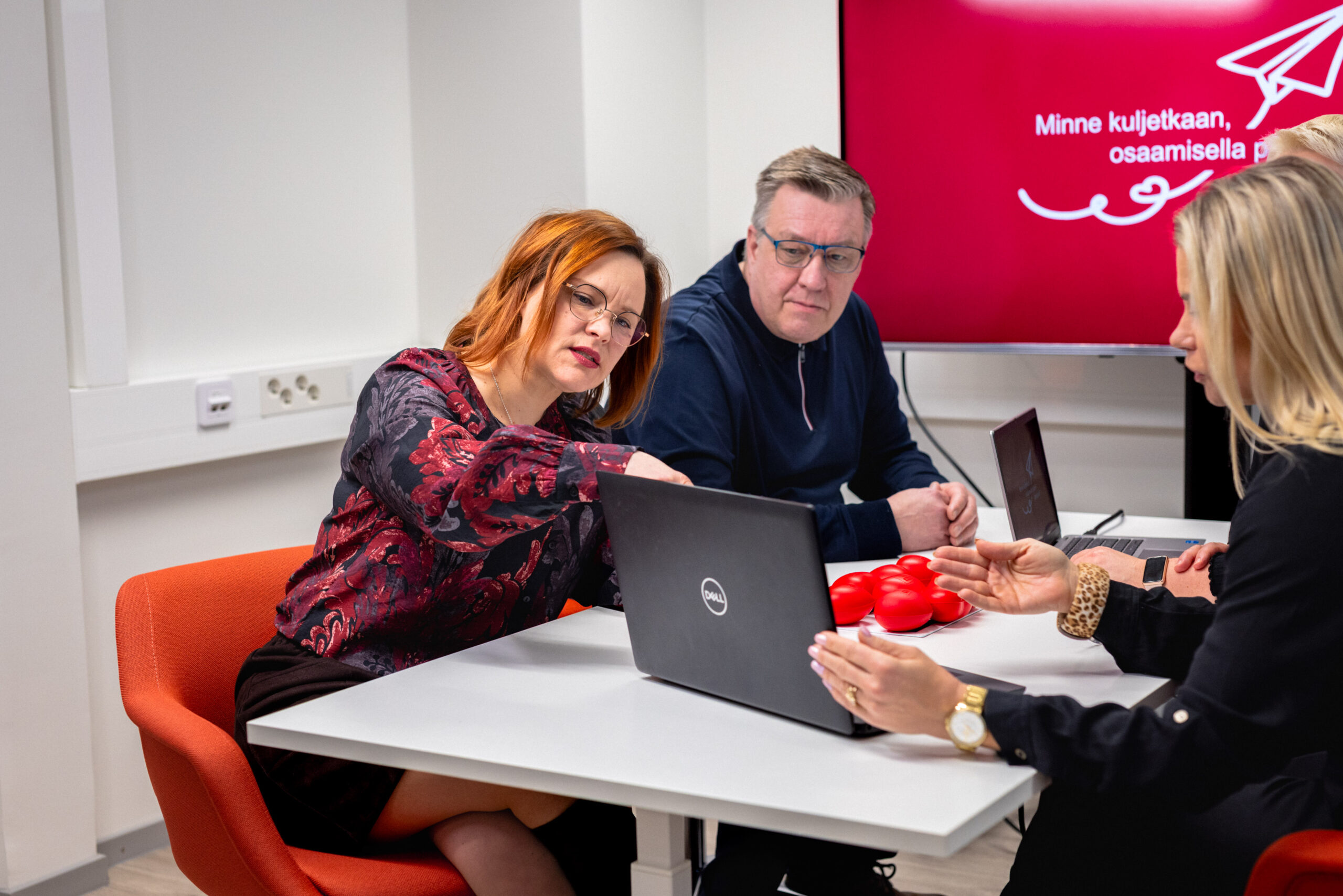 Neljä henkilöä istuu kokouspöydän ääressä ja tutkivat kannettavalta tietokoneelta jotain.