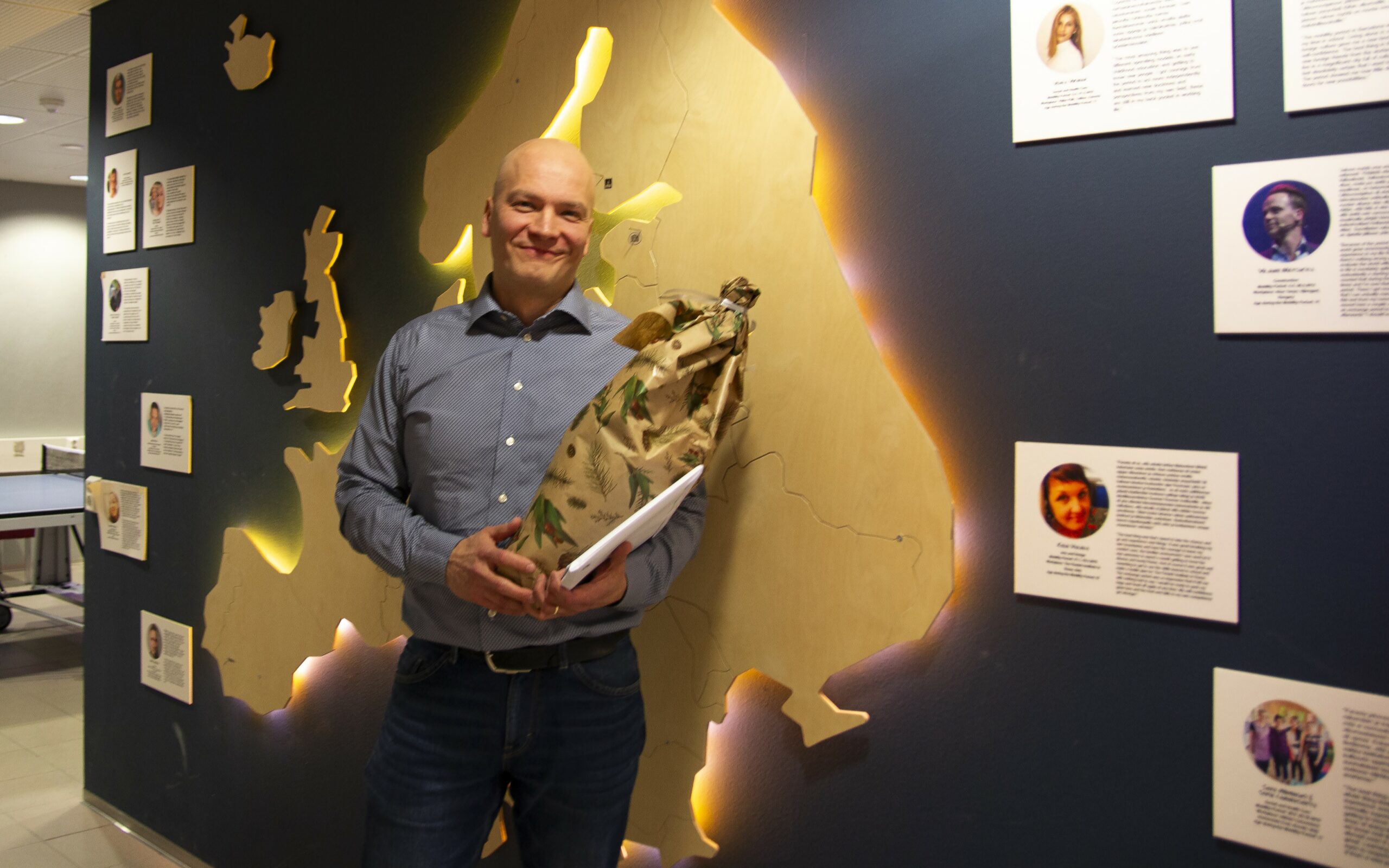 Valkeakosken ammattiopisto, VAAO, Henkilö seisoo Karttaseinän edessä kukkapaketti kädessään ja hymyilee kameralle