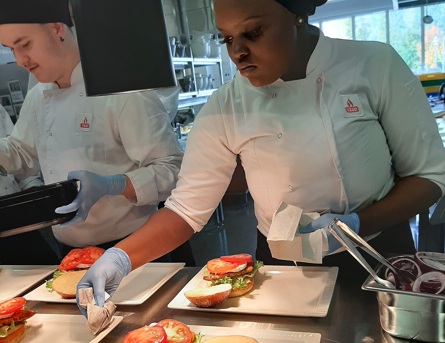 Valkeakosken ammattiopisto, VAAO, kaksi kokki-opiskelijaa valmistelee hampurilaisannoksia lautasille tarjoilua varten.