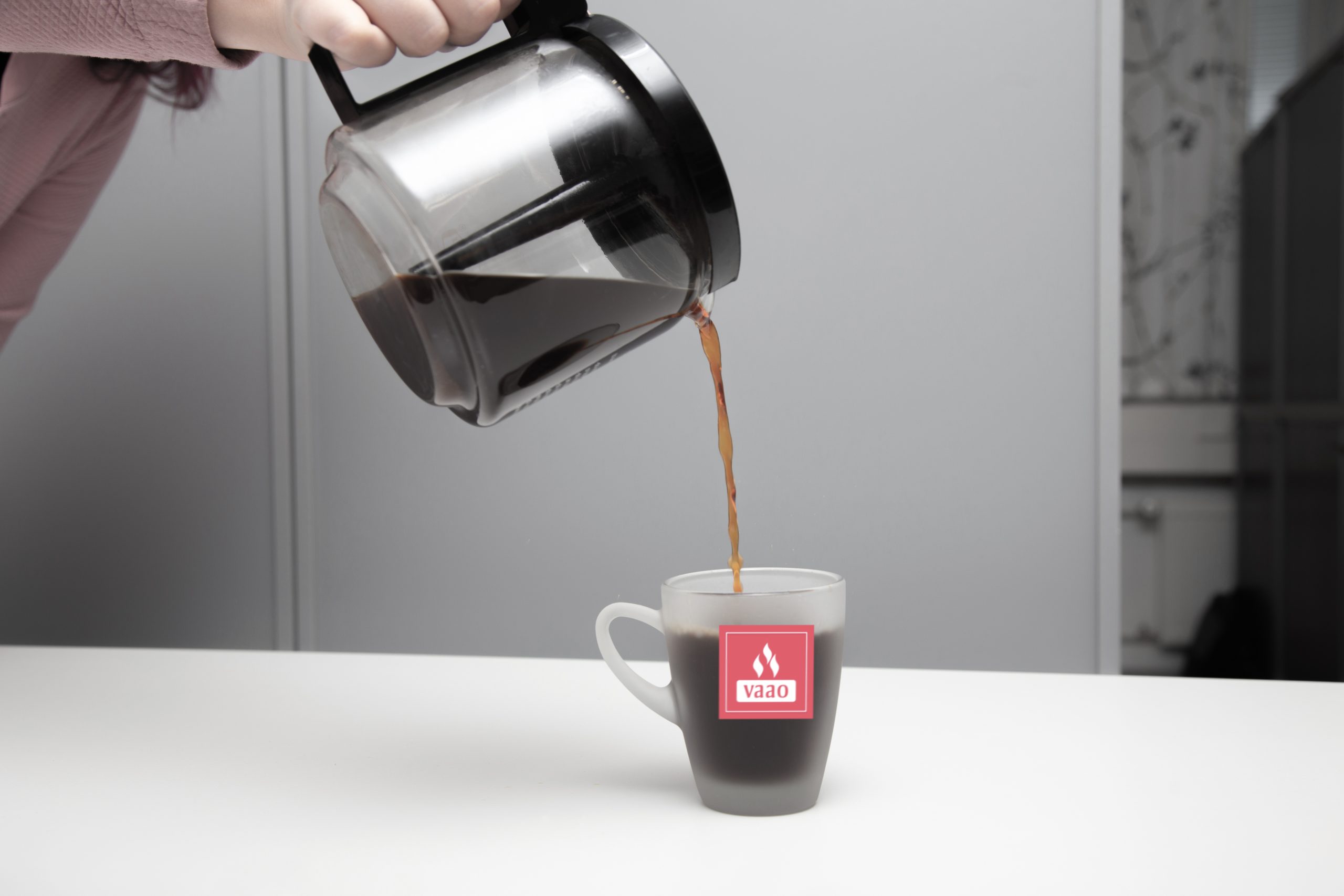 Valkeakosken ammattiopisto, VAAO. Läpinäkyvästä kahvipannusta kaadetaan kahvia pöydällä olevaan läpinäkyvään mukiin. Mukissa on VAAOn punainen logo.